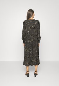 Frau mit langen braunen Haaren, die ein schwarzes Midikleid mit Blumen- und Punkte-Muster sowie schwarze Blockabsatzsandalen trägt und mit dem Rücken zur Kamera steht.