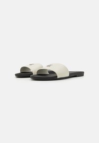Sandales en plastique avec un dessus synthétique crème, à bout ouvert, et une semelle en caoutchouc noire, présentant un détail de logo subtil à l'avant.