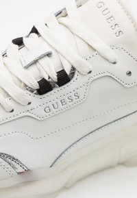 Zapatilla blanca de cuero con detalles texturizados, cordones en crema y negro, marca visible y suela de goma semitransparente.