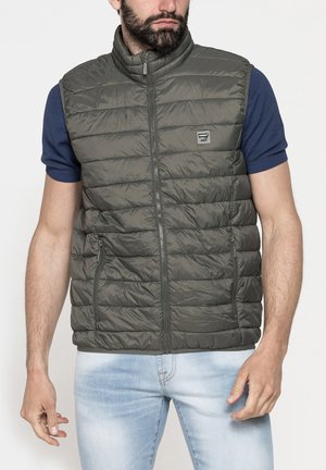 Uomo che indossa un gilet trapuntato senza maniche verde oliva sopra una camicia a maniche corte blu navy e jeans azzurri, in piedi davanti a uno sfondo semplice.