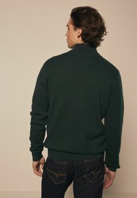 Maglione lavorato a maglia verde scuro con colletto alto e polsini a coste, indossato sopra jeans blu scuro con cuciture a contrasto e tasche posteriori.