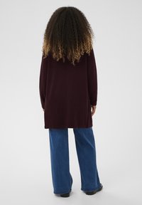 Bordowy sweter z długim rękawem o luźnym kroju, z zaokrąglonym dołem, w połączeniu z niebieskimi, szerokimi spodniami dżinsowymi i czarnymi butami. Widoczne kręcone włosy.