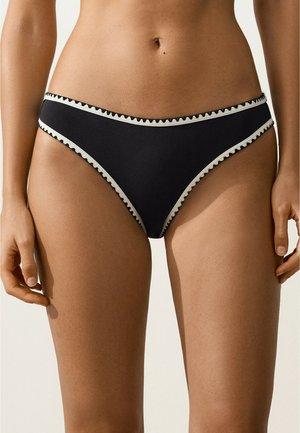 Kobieta nosząca czarne bikini z białym falowanym obszyciem, stojąca na jednolitym jasnym tle.