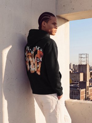 Un homme portant un sweat à capuche noir à motif graphique et un pantalon cargo blanc s’appuie contre un mur en béton sur un toit et contemple la ville.