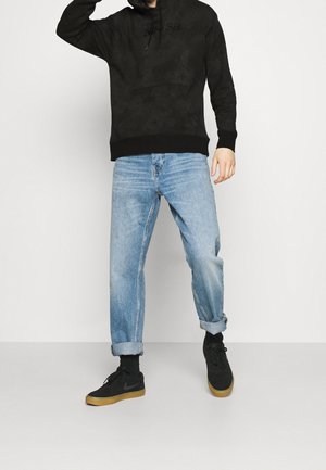 Schwarzer Hoodie mit einer Fronttasche und Logo, kombiniert mit hellblauen, hochgekrempelten Jeans aus Denim und schwarzen Sneakern mit Gummisohlen.