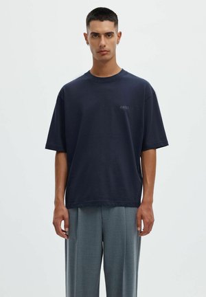 Ung mand med kort hår iført en mørk oversized t-shirt og løse grå bukser, stående mod en ensfarvet lys baggrund.