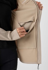 Personne tenant une veste beige texturée ouverte pour montrer la poche intérieure et les détails du tissu sur un fond neutre.