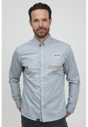 Camisa de botones azul claro con detalles bordados en el frente. Presenta un collar clásico y mangas largas, hecha de un tejido suave.