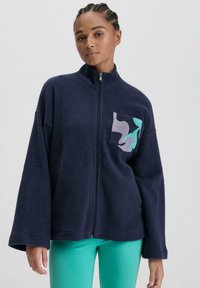 Marineblaues Fleece-Reißverschlussjacke mit lockerer Passform. Verfügt über eine Fronttasche mit einem bunten abstrakten Design in Lila und Türkis.