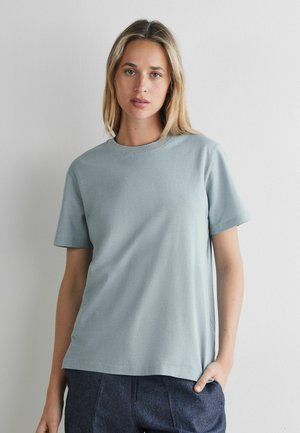 Kvinde med blondt hår iført en lyseblå kortærmet skjorte og mørkeblå bukser, stående mod en ensfarvet hvid baggrund.