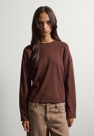 Donna con capelli lunghi e scuri che indossa una maglia marrone a maniche lunghe e pantaloni beige, in piedi davanti a uno sfondo chiaro e uniforme.