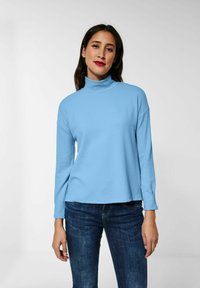 Maglione a collo alto di colore blu chiaro con un motivo testurizzato, maniche lunghe e vestibilità rilassata. Indossato con jeans di colore blu scuro.