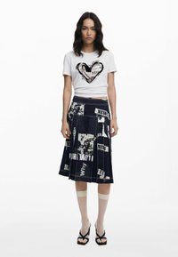 Weißes, kurz geschnittenes T-Shirt mit Herzgrafik, kombiniert mit einem marineblauen, plissierten Rock im Zeitungsdruck und schwarzen, geflochtenen Sandalen.