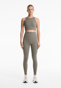 Mlada žena stoji okrenuta prema naprijed, obučena u usklađene maslinasto zelene tajice visokog struka i bezrukavi crop top, s bijelim tenisicama.