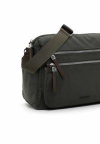Borsa in nylon verde scuro con molteplici tasche con zip, dettagli in pelle marrone e una tracolla regolabile. Texture liscia con una forma strutturata.