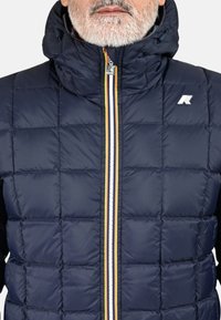 Veste matelassée bleu marine avec fermeture éclair à l'avant, détail de bandes jaunes et blanches, capuche, et texture matelassée. Logo présent sur la partie supérieure de la poitrine.