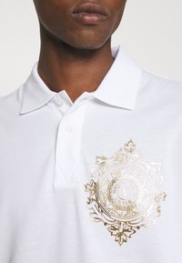 Weißes Poloshirt aus strukturiertem Stoff mit einem goldenen bestickten Emblem auf der linken Brust. Sichtbarer Zwei-Knopf-Verschluss und Krageninformation.