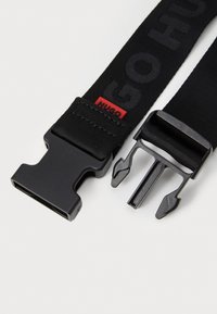 Ceinture noire en tissu avec boucle en métal au design élégant. Comprend une étiquette logo rouge. Matériau durable avec une finition texturée.