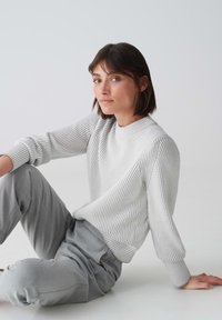 Hellgrauer gerippter Pullover mit hohem Kragen, lockerem Schnitt und langen, leicht puffigen Ärmeln. Kombiniert mit lockeren grauen Hosen. Minimalistisches Design.