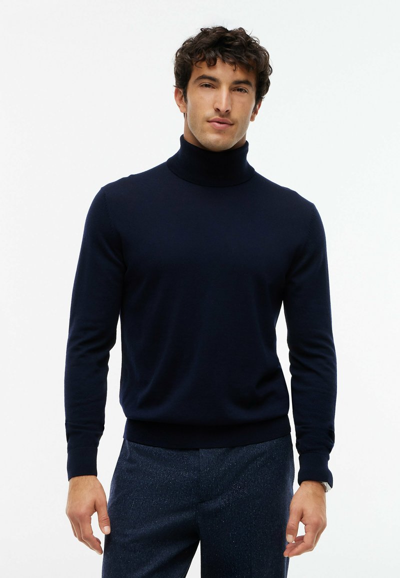 Homme aux cheveux courts et bouclés portant un pull à col roulé bleu marine foncé et un pantalon bleu foncé sur un fond blanc uni.