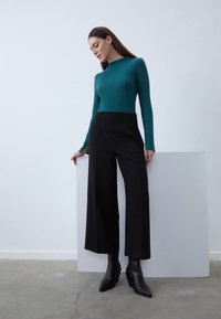 Naine türkiissinises pika varrukaga pluusis ja mustades laiade säärtega pükstes toetub minimalistlikus toas, millel on hall põrand, valgele paneelile.