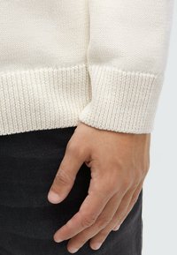 Cremefarbener, gestrickter Pullover mit geripptem Saum und Bündchen, der eine weiche Textur aufweist. Eine Hand ruht im Vordergrund auf schwarzem Denimstoff.