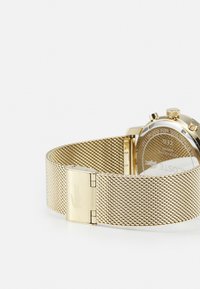 Montre en mailles dorées avec un boîtier rond et un design minimaliste ; présente une fermeture à boucle et un logo gravé sur le bracelet.