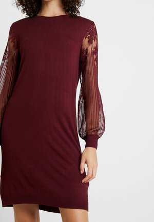 Woman wearing a burgundy knit dress with sheer, floral lace balloon sleeves and ribbed cuffs, standing against a plain light background.