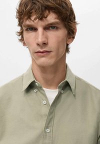 Camisa de botones verde claro con cuello, de textura lisa y botones pequeños. Lleva una camiseta blanca debajo, mostrando un estilo casual.