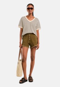 Juostuota v-kaklo marškinėliai smėlio ir juodos spalvos, alyvuogių žalumo šortai su trimis mygtukais priekyje ir pakraščiu su frangėmis, derinami su sandalais ir nertiniu krepšiu.