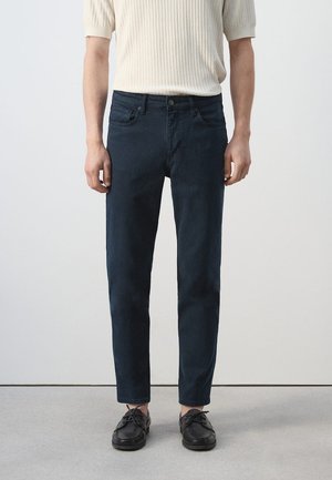 Mann trägt dunkelblaue Slim-Fit-Jeans, cremefarbenen kurzärmeligen Rippsweater und schwarze Schnürschuhe, steht vor einfachem neutralem Hintergrund.