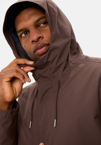 Giacca marrone con cappuccio realizzata in materiale liscio e resistente all'acqua; presenta un cordoncino e inserti metallici all'apertura del cappuccio.