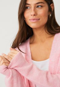 Cardigan rose pâle en matière duveteuse, avec des poignets côtelés et un design léger. Porté sur un débardeur blanc.