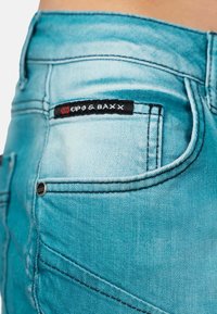 Jean en denim bleu clair avec une texture délavée, arborant une étiquette noire avec l'inscription "GPO & BAXX" sur la poche arrière. Détails de couture visibles.