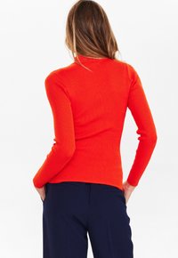 Gerippter Langarmoberteil in leuchtendem Orange, mit einem taillierten Schnitt und Rundhalsausschnitt. Getragen mit dunkelblauen Hosen.