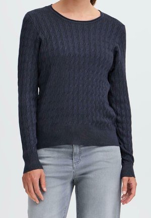 Vrouw die een marineblauwe kabelgebreide trui met ronde hals en lange mouwen draagt, gecombineerd met lichtgrijze jeans.