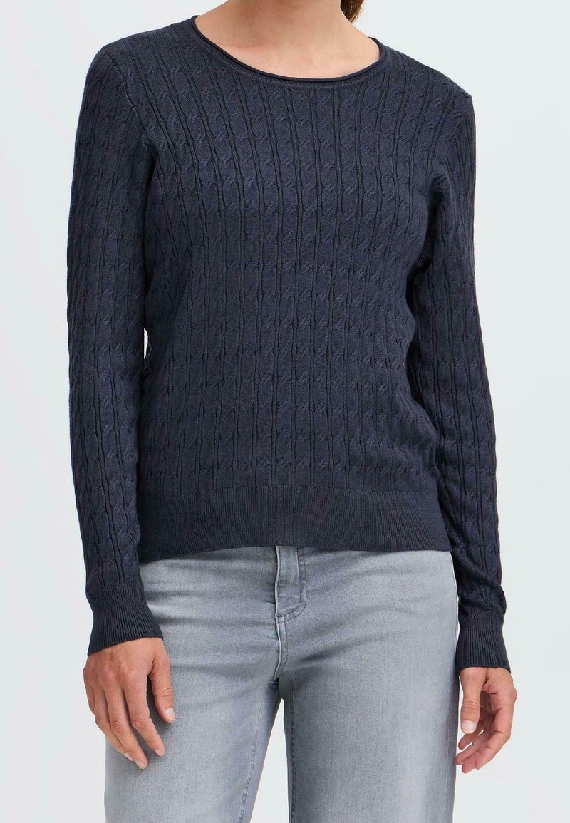 Femme portant un pull en maille torsadée bleu marine avec col rond et manches longues, associé à un jean gris clair.