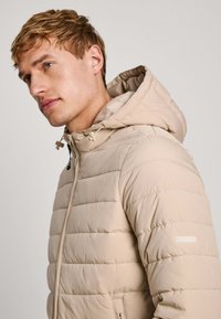 Chaqueta acolchada beige con capucha, diseño acolchado, detalles de cordón, cierre de cremallera y bolsillos laterales. Ligera y con una textura suave.