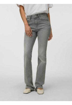 Person iført højtaljede grå jeans med lige ben, hvid top og lysebeige spidse sko mod en ensfarvet baggrund.