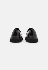 Sorte lakk lær sko med rund tå, tykk svart gummisåle og lav hæl. Har minimalistisk design og stikkingdetaljer.