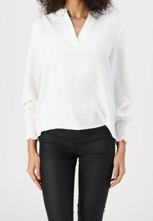 Femme portant un chemisier blanc à manches longues avec un col en V et des poignets froncés, associé à un pantalon noir serré, debout devant un fond uni.
