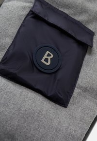 Grijze stof met een marineblauwe zak met klep, ronde logo-detail en een gladde textuur. Het algehele ontwerp is functioneel en praktisch.