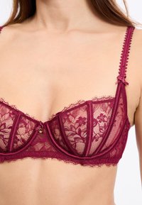 Bordowy koronkowy biustonosz z fiszbinami, kwiatowym wzorem, falistymi krawędziami i regulowanymi ramiączkami. Posiada centralną kokardkę i złote elementy.