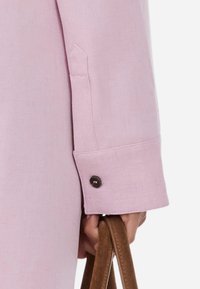 Μανίκι από ανοιχτό ροζ σακάκι με λεπτομέρεια κουμπιού και ραμμένο μανίκι. Ο υφασμάτινος ιστός δημιουργεί μια διακριτική λάμψη, σε συνδυασμό με μια καφέ λαβή τσάντας.