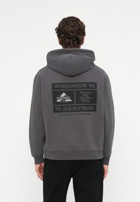 Γκρι φούτερ με μεγάλο τύπωμα στην πλάτη που γράφει "WORLDWIDE '85" και "tommy jeans" σε μαύρο χρώμα. Περιλαμβάνει κουκούλα και πλεκτούς μανικιών.