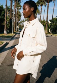 Jauns cilvēks gaiši krēmkrāsas pārāk lielā krekla krekla stāv saulainā ielā ar palmu kokiem.