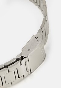 Bracelet de montre en acier inoxydable, texture brossée, couleur argent, design à maillons avec une boucle comportant une marque gravée et de petits trous pour le réglage.