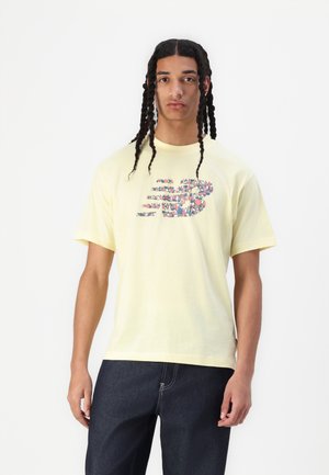Camiseta de algodón amarilla con mangas cortas que presenta un diseño gráfico de logotipo multicolor en el frente. Llevada con pantalones de mezclilla oscuros.