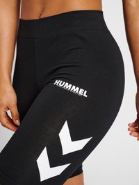 Close-up van zwarte nauwsluitende korte broek met wit Hummel-logo en chevron-patronen op de bovenkant van het bovenbeen van een persoon met zichtbare handen.