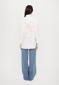 Witte shirt met een kraag, met een doorzichtige achterkant en lichte palmbomenprints. Gecombineerd met wijde, lichtblauwe jeans.